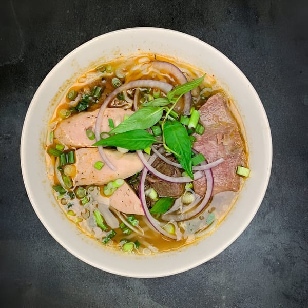 A bowl of Vietnamese pho with beef, pork, noodles, red onions, and basil on a dark surface.
