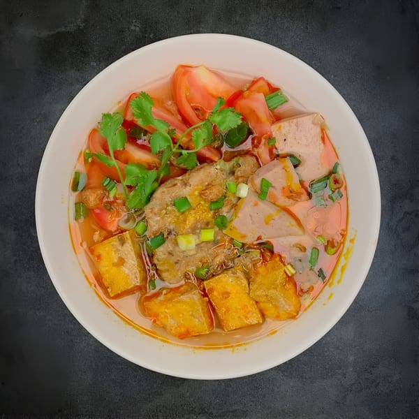 A vibrant Vietnamese soup with tomatoes, tofu, and meat in a white bowl.